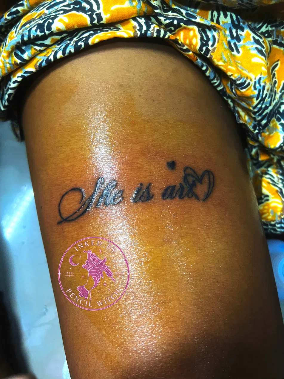 Cursive script tattoo reading 'she is art' with a small heart, on the thigh
